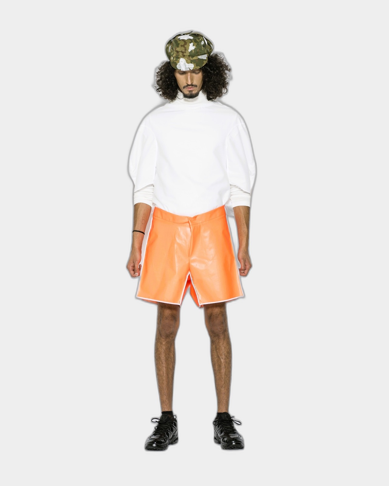 White turtleneck top with neon-orange coated leather shorts and camo cap